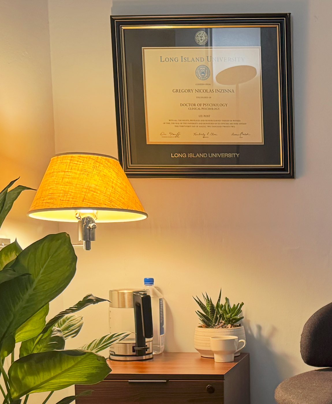 Corner of Dr. Inzinna's Upper East Side office with a warm reading lamp, a framed Doctor of Psychology diploma from Long Island University, plants and a small tea station